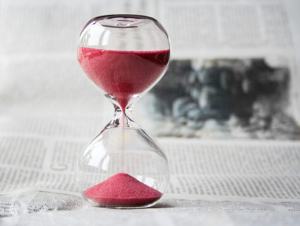 hourglass time hours sand 39396 39396 Close-up of a transparent hourglass with pink sand flowing, placed on a newspaper background.
