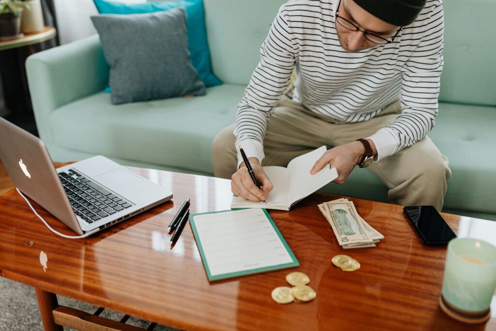 pexels photo 6329021 6329021 A man in a striped shirt sitting at a wooden table, writing in a notebook with money and a laptop nearby.