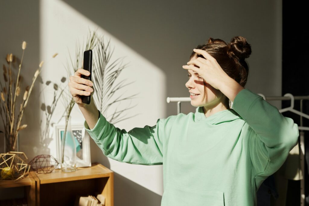 A young girl in a green hoodie takes a selfie indoors, enjoying the sunlight.
