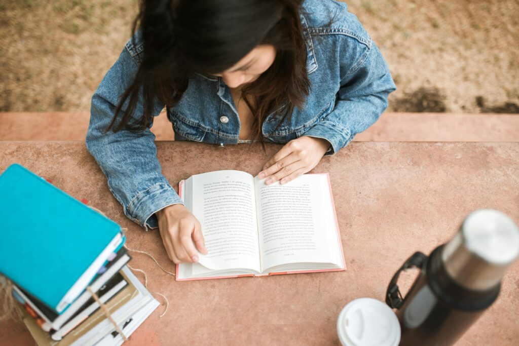 pexels photo 5530681 5530681 Overhead view of a woman reading a book outdoors on a sunny day, wearing a denim jacket.